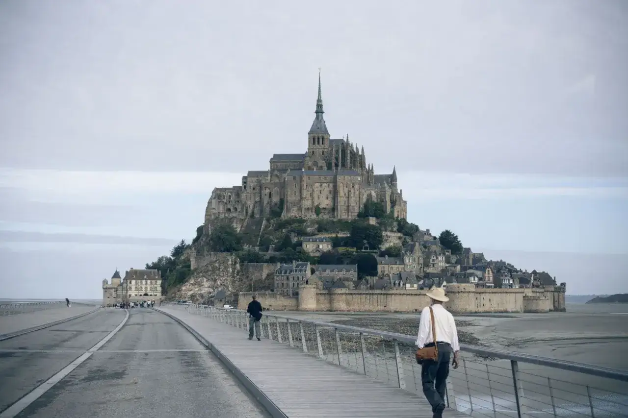 En man med hatt går på en gångbro mot Mont Saint-Michel, en av Frankrikes mest ikoniska sevärdheter.