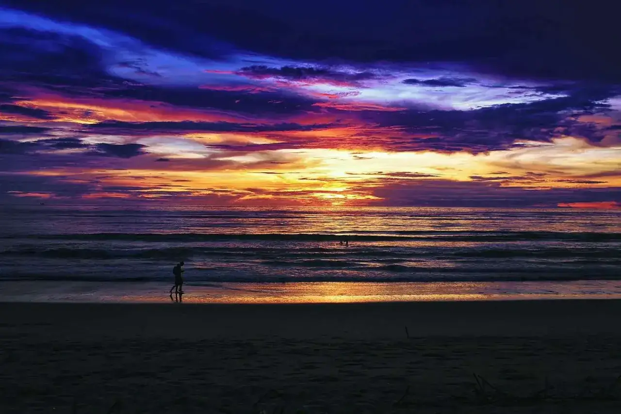 Ensam person promenerar på stranden vid en dramatisk solnedgång i Thailand, Khao Lak. Himlen exploderar i lila och orange.