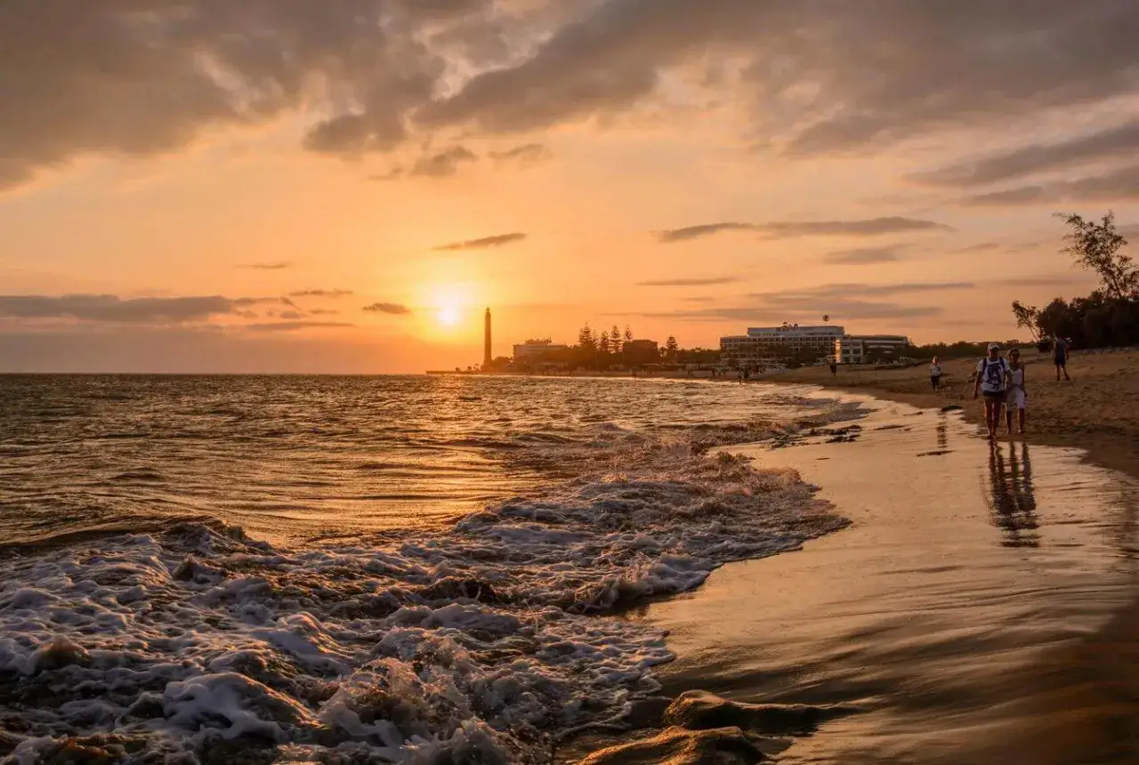 Solnedg&aring;ng &ouml;ver Dunas de Maspalomas med v&aring;gor som sk&ouml;ljer in mot stranden. En fyr och hotell syns i bakgrunden.