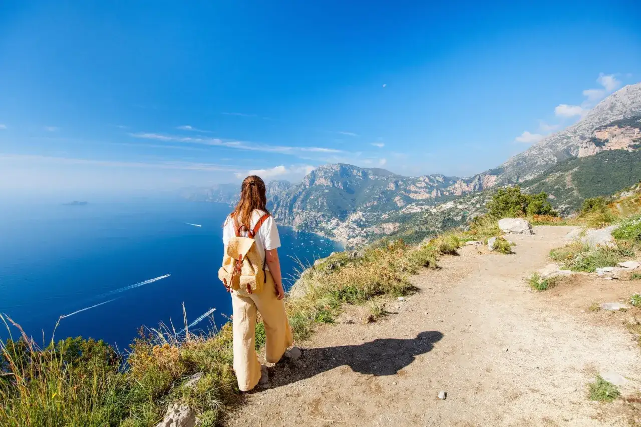En kvinna med ryggs&auml;ck njuter av utsikten &ouml;ver en kuststad vid havet under en vandring i Italien.