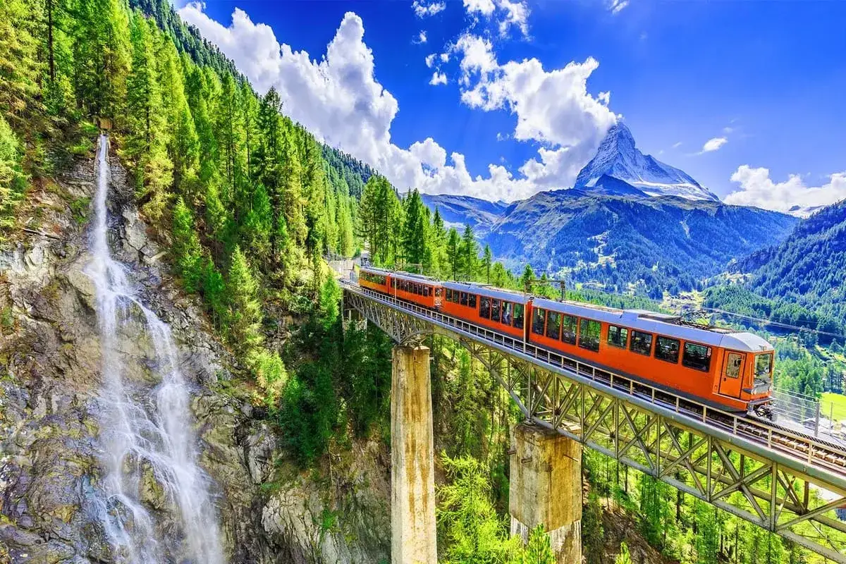 Ett orange tåg korsar en bro över en forsande vattenfall i Schweiz, med majestätiska berg och en blå himmel i bakgrunden.