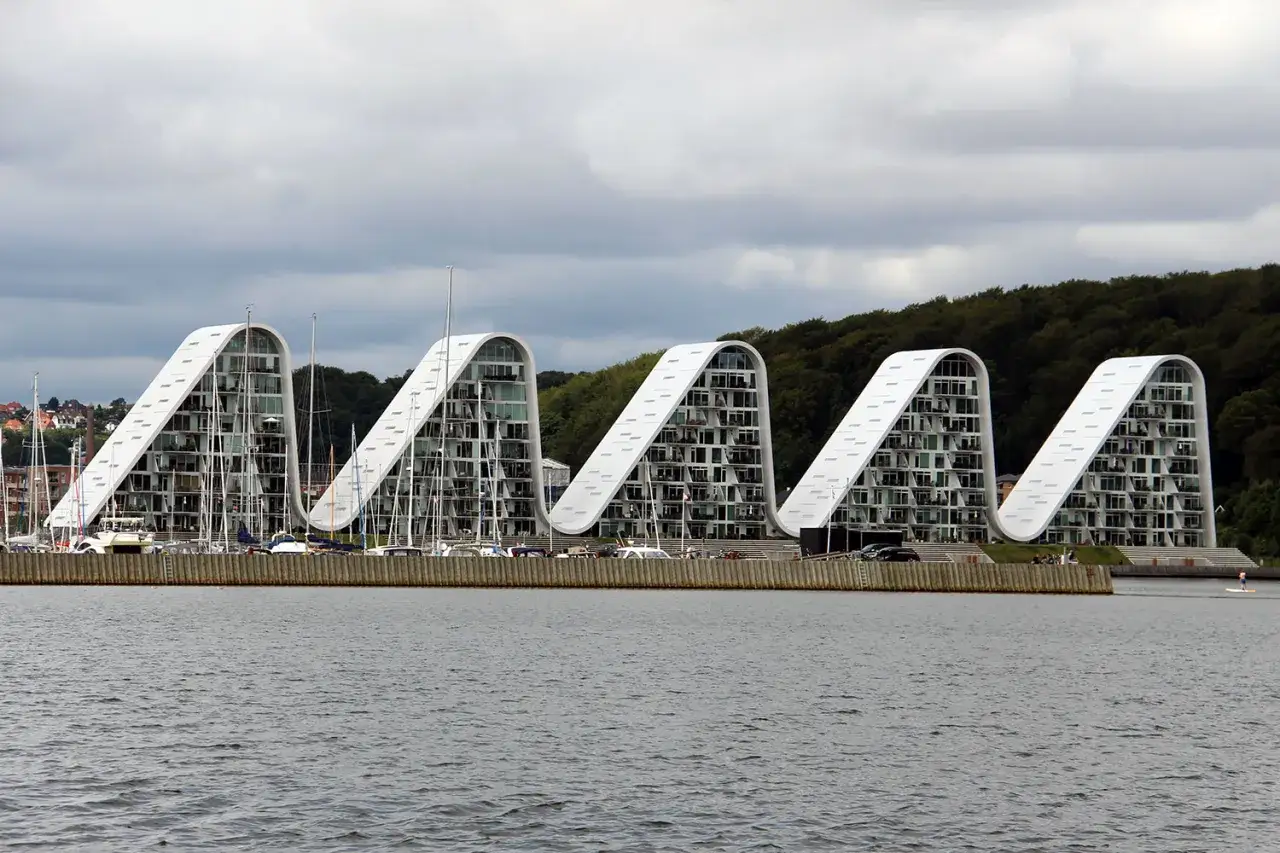 Vågformade byggnader vid vattnet i Vejle, Danmark, med segelbåtar i hamnen.
