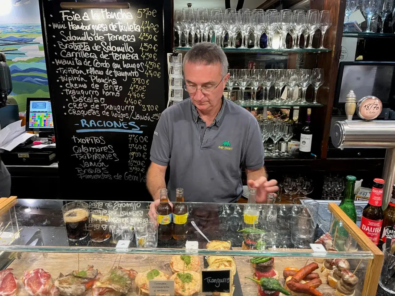 Man vid en bardisk i San Sebastián, med en meny med tapas och raciones på svarta tavlan.