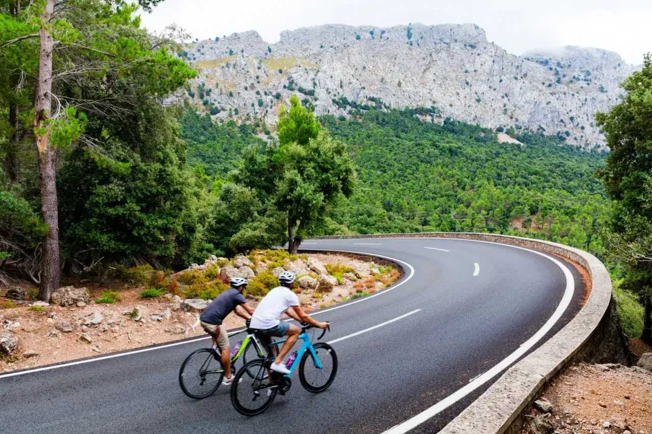 Tv&aring; m&auml;n cyklar p&aring; en slingrig v&auml;g p&aring; Mallorca, med gr&ouml;nskande berg i bakgrunden.