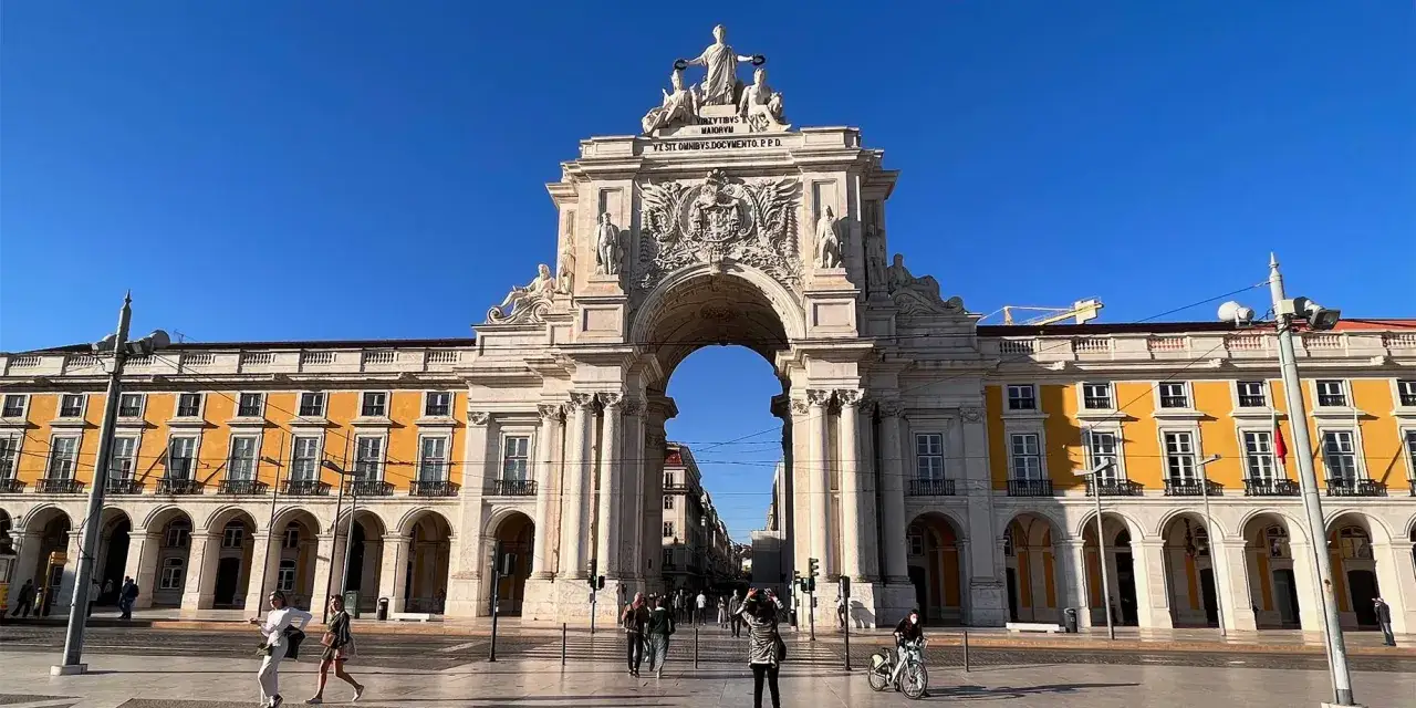 En solig dag i Lissabon gamla stan. En imponerande triumfbåge dominerar torget, med folk som promenerar och cyklar.
