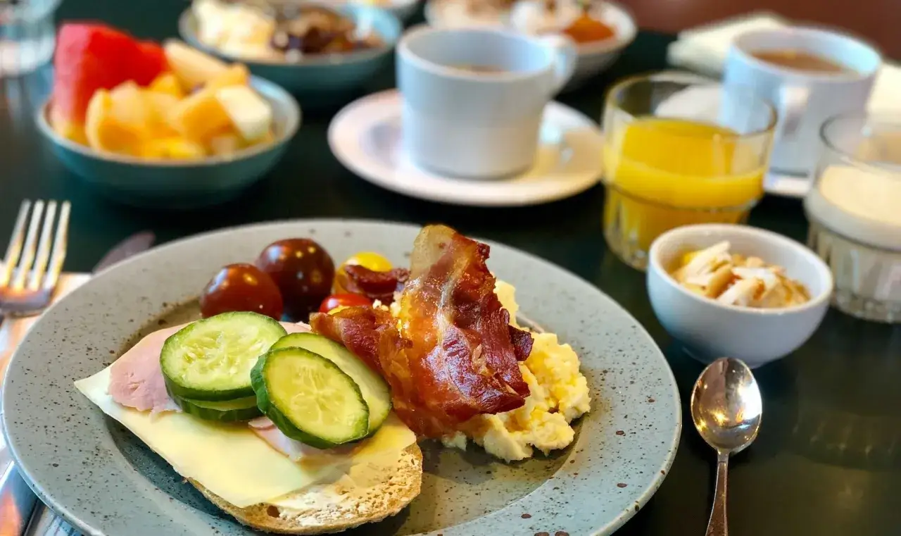 En härlig continental frukost med äggröra, bacon, smörgås med ost och skinka, tomater, gurka, frukt och juice.