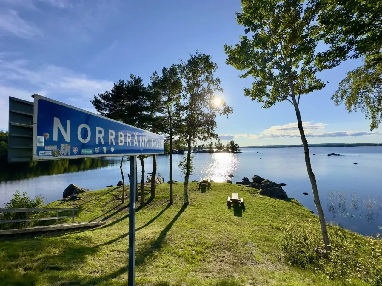 Vacker rastplats vid Norrbränningen, E4 norrut. Solen skiner över sjön, med picknickbord och träd.