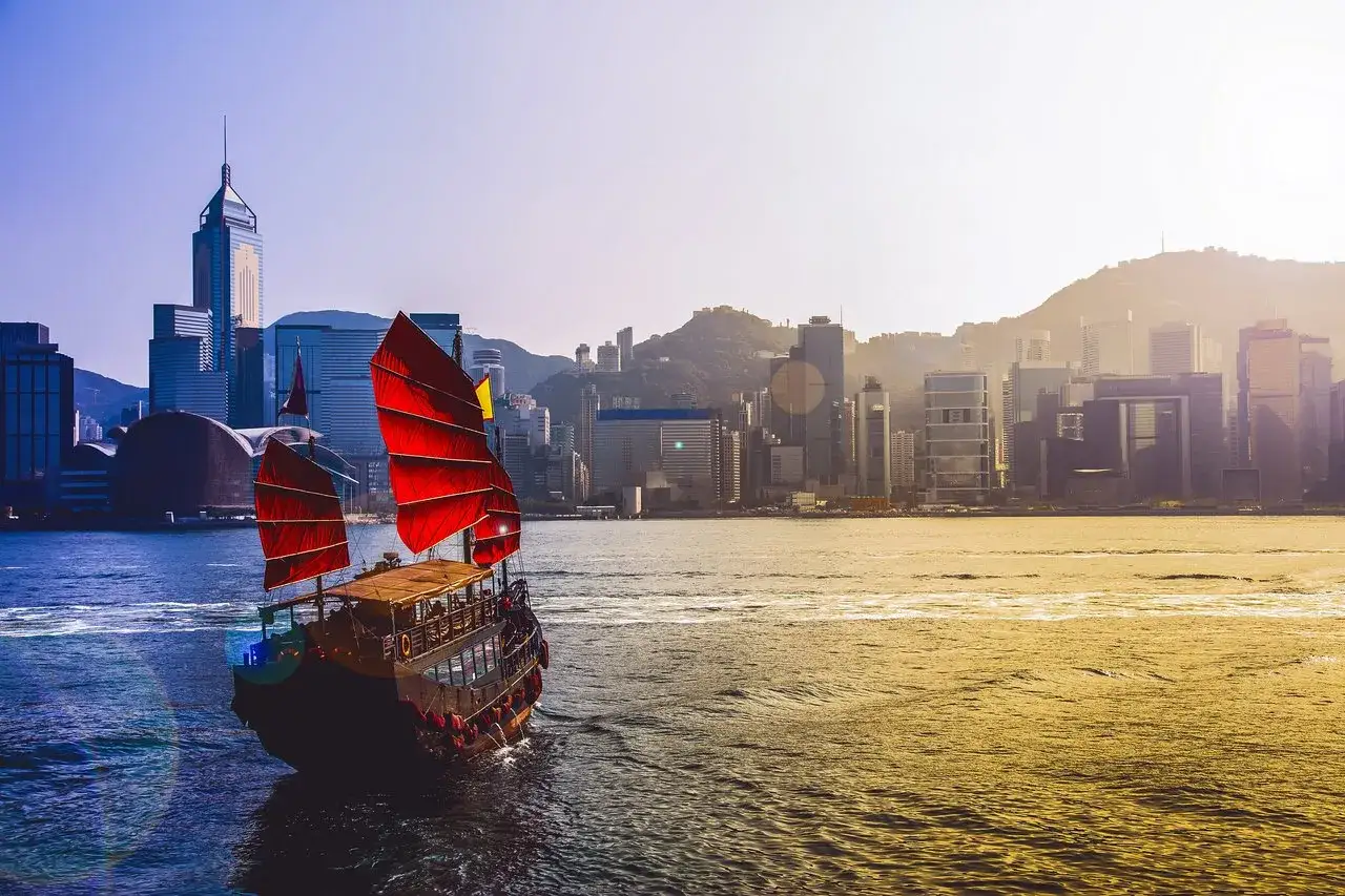 En traditionell kinesisk junk med röda segel glider fram på vattnet mot Hong Kongs imponerande skyline.