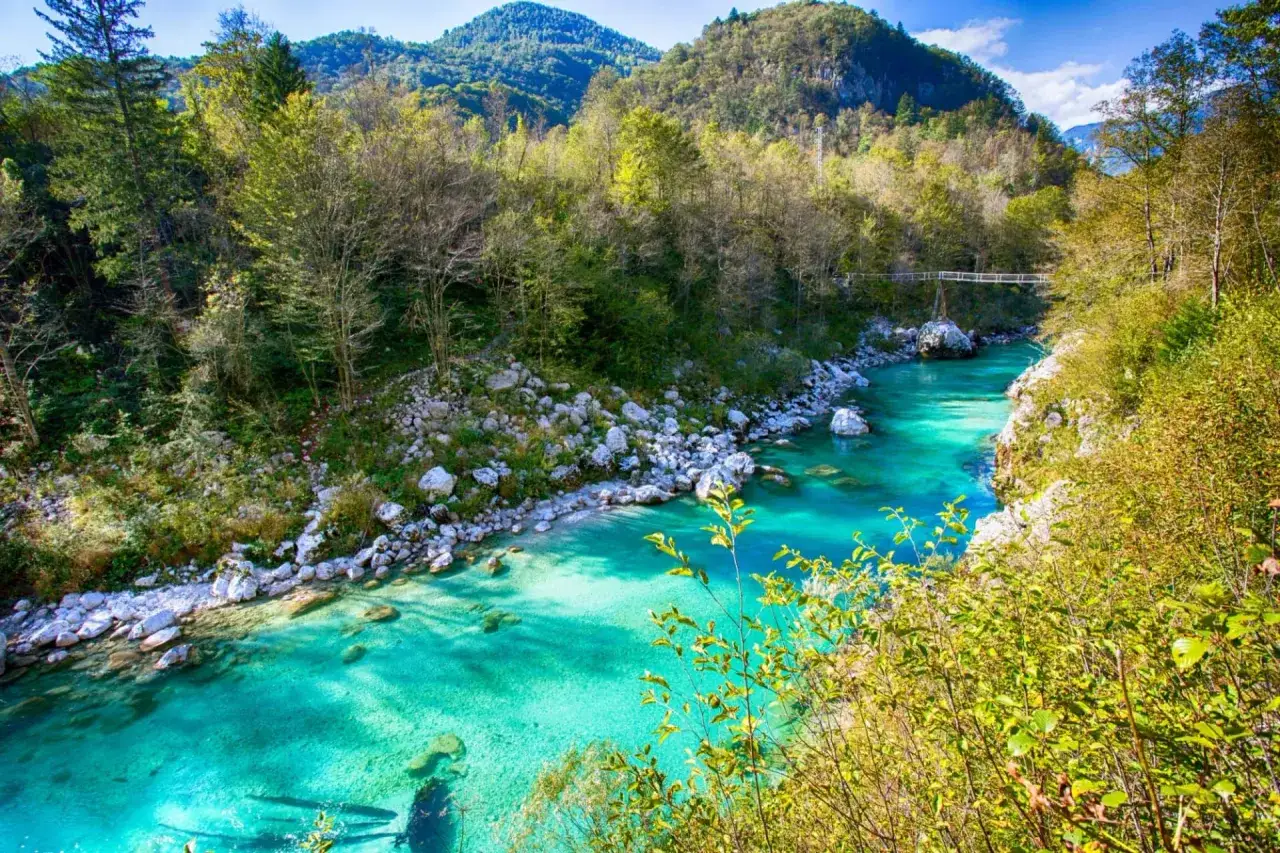 Vandra i Slovenien längs en turkos flod kantad av stenar och grönskande berg. En bro syns i fjärran.