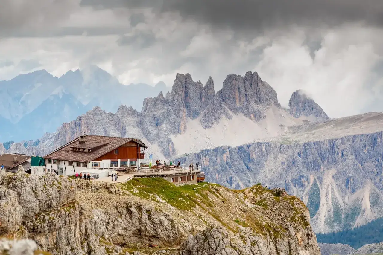 En fjällstuga med terrass och gäster, belägen på en klippavsats med dramatiska berg i bakgrunden. Perfekt för en paus under en dolomiterna vandring.