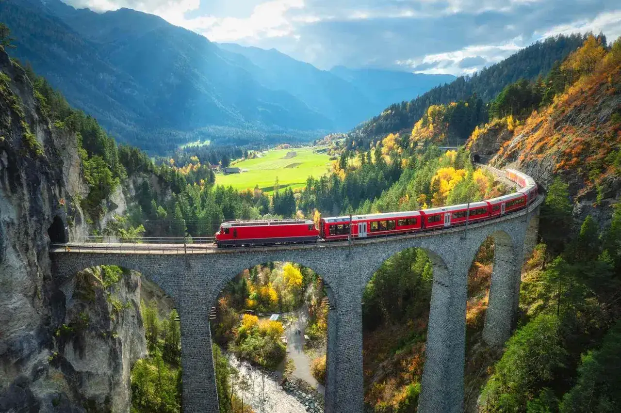 Bernina Express slingrar sig över en stenbro genom en höstfärgad dal med gröna fält och blå berg i bakgrunden.