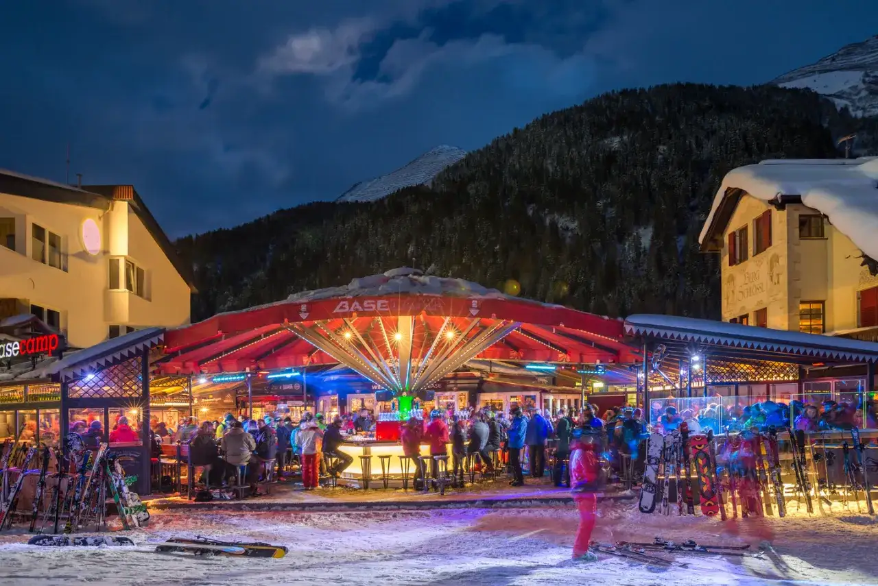 Livligt ställe för st anton afterski. Folk minglar vid baren under en upplyst röd parasoll. Skidor och snowboards står uppradade i snön.