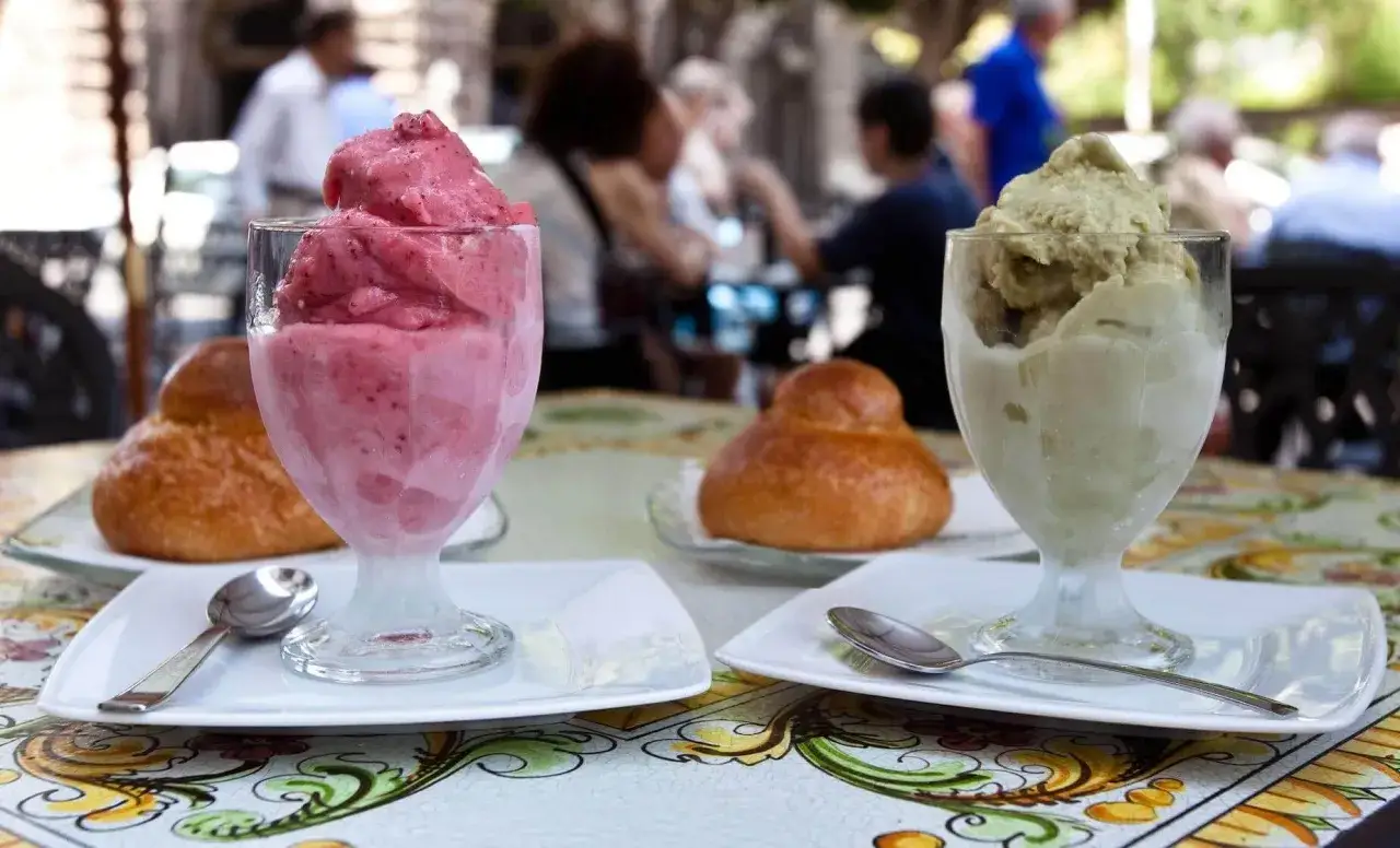 Två glas med glass, en rosa och en grön, står på ett bord med mönstrad duk. I bakgrunden syns människor på en uteservering, kanske på en av alla mysiga restauranger i Aten.