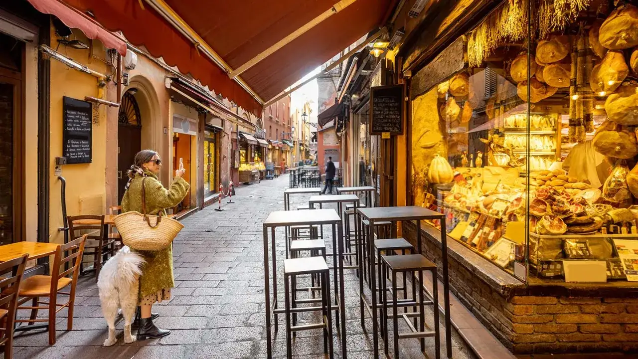 En kvinna med hund tar foto på en charmig gata i Bologna, Italien, kantad av delikatesser.