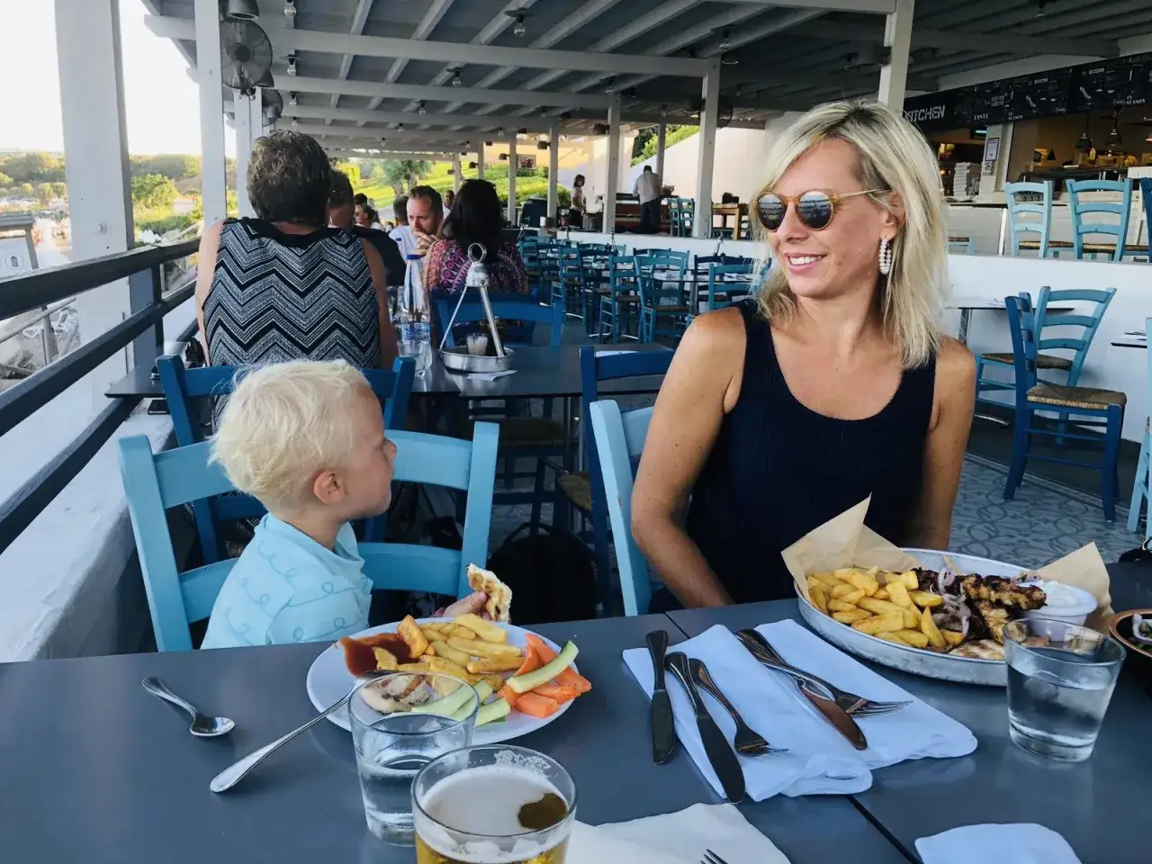 Familj njuter av en måltid på Costas Kitchen Kallithea. En kvinna ler, en pojke äter, och maten ser utsökt ut.