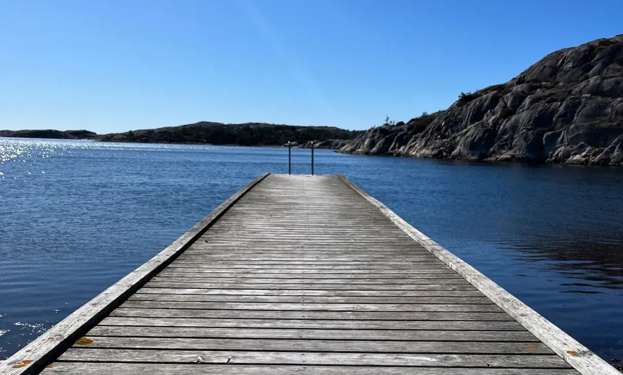 Träbrygga leder ut mot det glittrande vattnet vid Grebbestad strand. Klippor och öar syns i bakgrunden under en klarblå himmel.