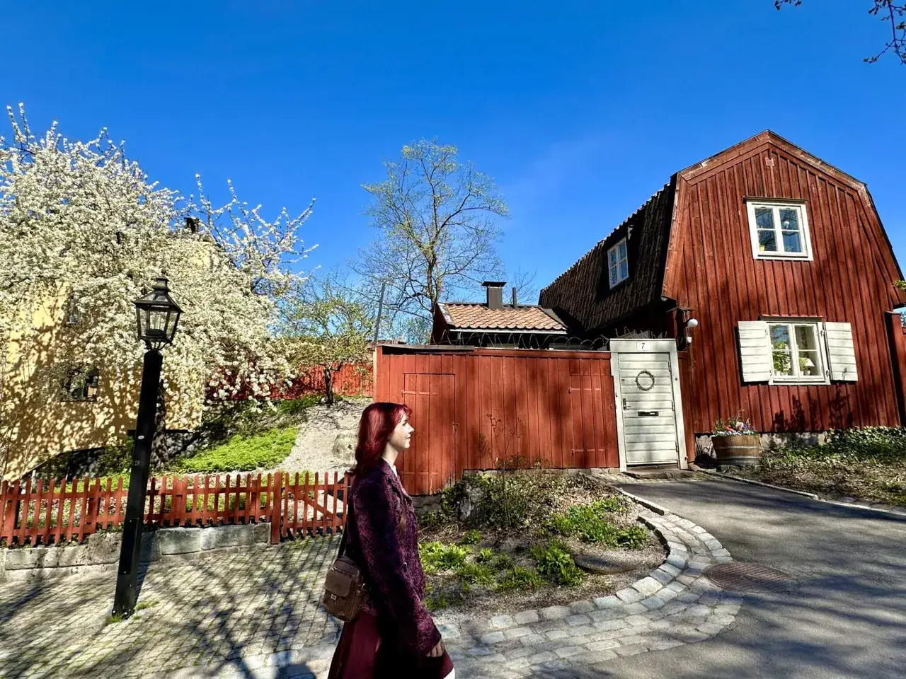 En kvinna med r&ouml;tt h&aring;r promenerar l&auml;ngs ett charmigt promenadstr&aring;k i Stockholm, f&ouml;rbi r&ouml;da tr&auml;hus och blommande tr&auml;d.