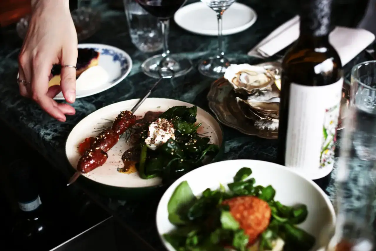 En hand sträcker sig efter en spettad köttbit på en tallrik med grönsaker. Bredvid finns ostron, vin och en sallad. En härlig måltid, kanske på Odenplan.
