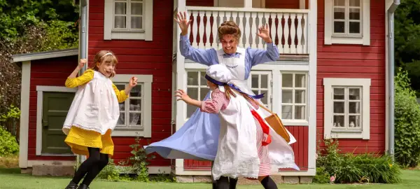 Barn leker vid Junibacken Madicken hus. En vuxen i blå klänning och två barn i tidsenliga kläder.