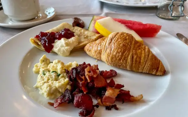 En härlig **continental frukost** med krämiga äggröror, knaprigt bacon, en croissant, pannkakor med sylt och grädde, samt färsk frukt.