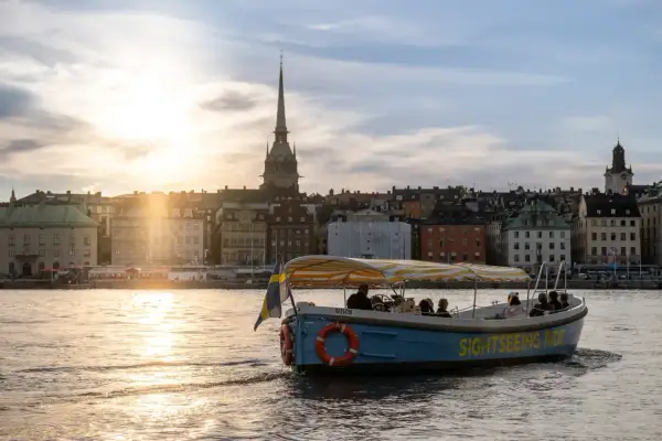 A blue sightseeing boat with a yellow striped canopy sails on water in Stockholm, with historic buildings and a church spire in the background at sunset.