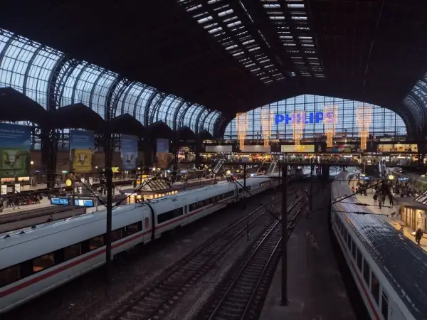 Tågstation med flera spår och vita tåg. En stor glas- och stålkonstruktion täcker taket. Philips-logotypen lyser i blått. Det är ett sj tåg till Hamburg.