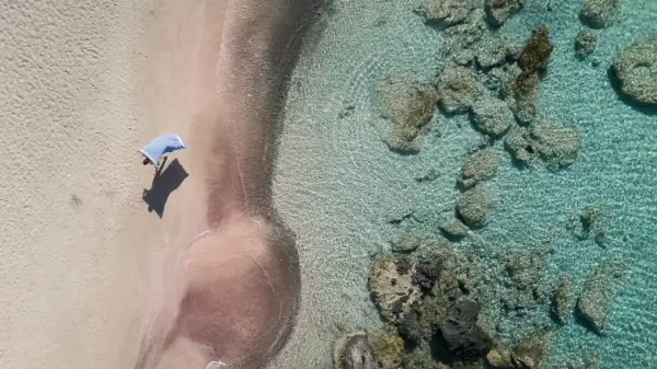 En person med en blå handduk på en rosa strand på Kreta. Klart turkost vatten med stenar.