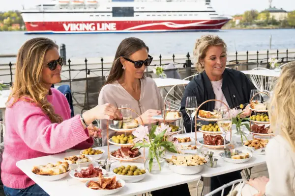Kvinnor njuter av mat och dryck vid ett bord på en sq restaurang med Viking Line-färja i bakgrunden.